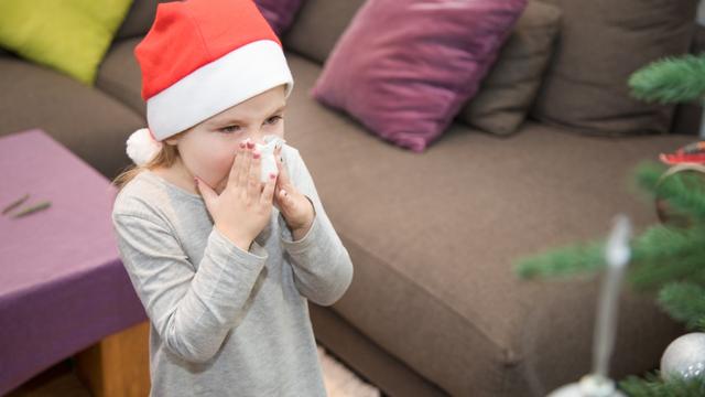 four years blonde cute girl with red Santa Claus hat, blowing her nose with white tissue next to Christmas tree at home
