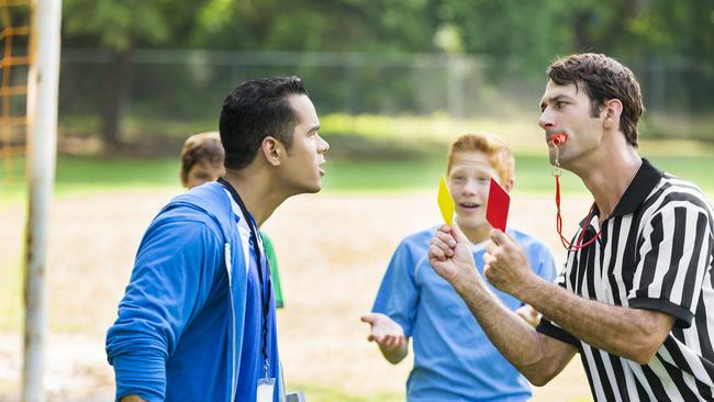 Mid adult Hispanic soccer coach argues with mid adult Caucasian referee about a penalty call. The referee has a whistle in his mouth and is showing the coach red and yellow cards. The players are in the background gesturing in unbelief. The coach is wearing a blue hoodie and the players are wearing blue jerseys. The playing field is in the background.  - Picture iStock