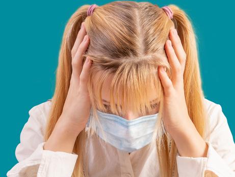 A schoolgirl in a mask on her face sits at a desk and clutching her head with her hands. Front view, place for text, Copy space, turquoise background.