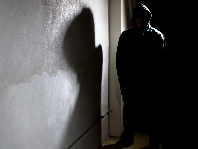 Photo of a hooded criminal stalking in the shadows of a dark street alley.  The hooded man is a silhouette and hiding in the dark.  The man is a criminal waiting to ambush victims.  The concrete walls provide copyspace.The photo depicts crime.  - istock