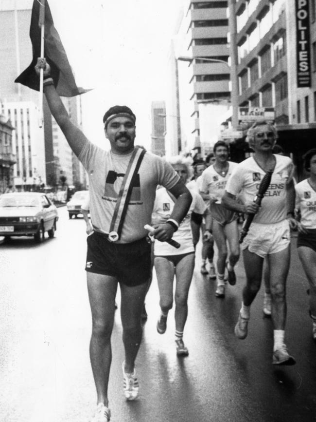 Mundine carries the Aboriginal flag as he runs alongside Commonwealth Games torch relay runner Ian Perrin as he runs down King William Street to the Adelaide Town Hall.