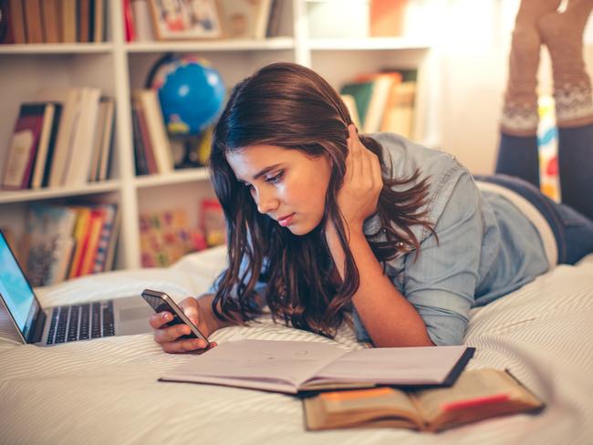 Teenage girl lying on bed in her room and studying. Using phone to check news or messages.  Evening or night with beautiful yellow lights lightning the scenes.