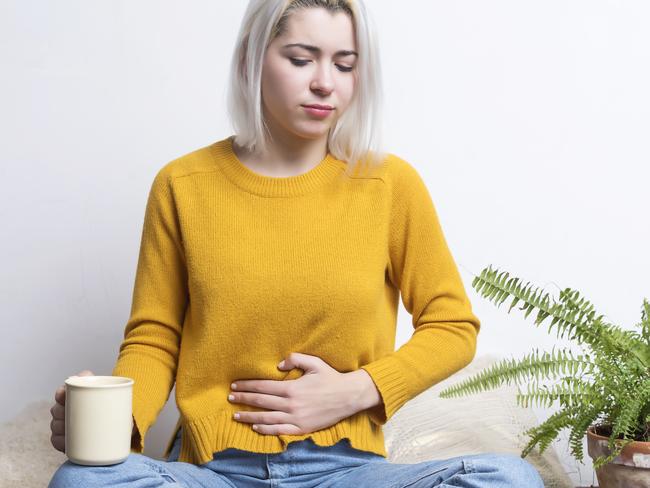 Young female sitting at home holding a cup of tea while putting her hand in her stomach