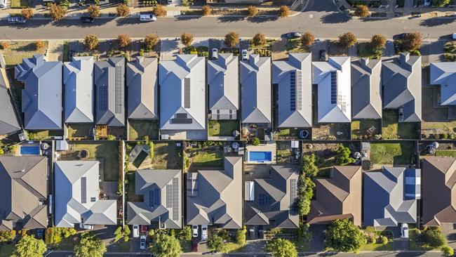 Aerial view directly above new outer suburban/semi-rural housing development with single-level housing between two streets with t-intersections and orange coloured street trees.  Mostly gray metal roofing, landscaped front and back yards, some solar panels, cars and motor vehicles parked in driveways and street, some street and parkland trees, water tanks in backyards, sheds and garages, swimming pools.  Mount Barker, South Australia; property investment housing money generic