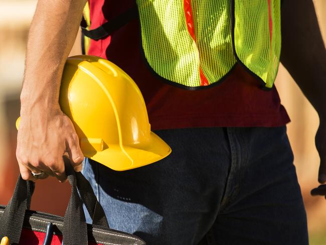 A construction worker busy working at a job site. He holds a tool box full of tools and a hard hat. Framed house, building in background. He is wearing a safety vest.