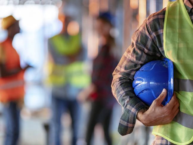 Man holding blue helmet close up. Construction man worker with office and people in background. Close up of a construction worker's hand holding working helmet.