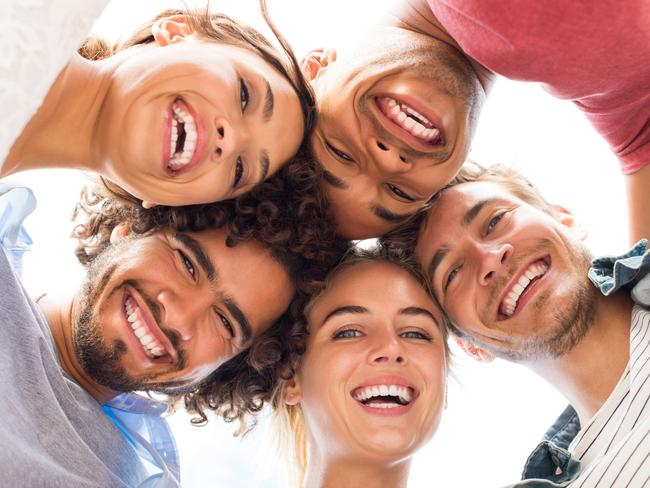 Directly below shot of young friends forming huddle. Low angle view of girls and guys with their head forming a circle. Portrait of young people looking at camera. Friendship and unity concept.