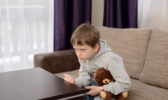 Annoyed 7 year old boy child sitting on the sofa and watching tv. Hugs his favorite teddy bear and changing channels by remote control.