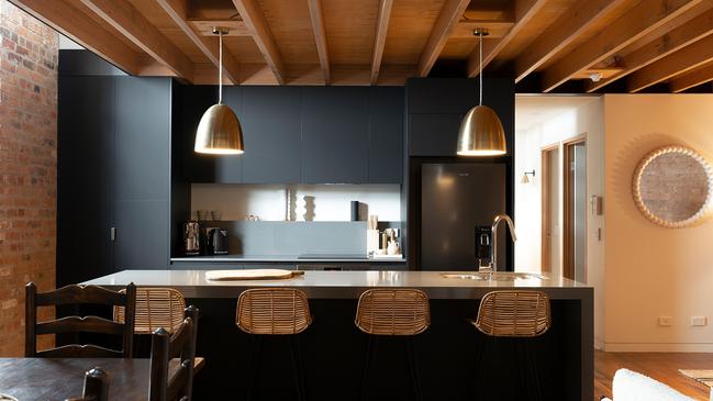 The Elizabeth Apartment’s sleek black, matt contemporary kitchen cabinetry melds beautifully with the brass light pendants, cane barstools and natural timbers. Picture: AARON JONES