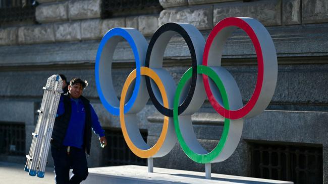 A man carrying a lader walks past the Olympic rings set at the entrance of Milan's city all on February 14, 2024 in Milan. Milan and Cortina will host the 2026 Winter Olympic Games. (Photo by GABRIEL BOUYS / AFP)