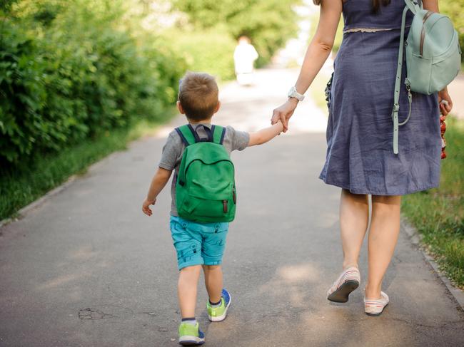 Back view of mother walking down the street with a little son with a backpack on sunny summer day