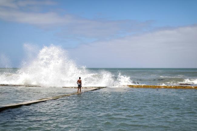 A man walks on a sea wall as waves spray water on Kalk Bay in Cape Town