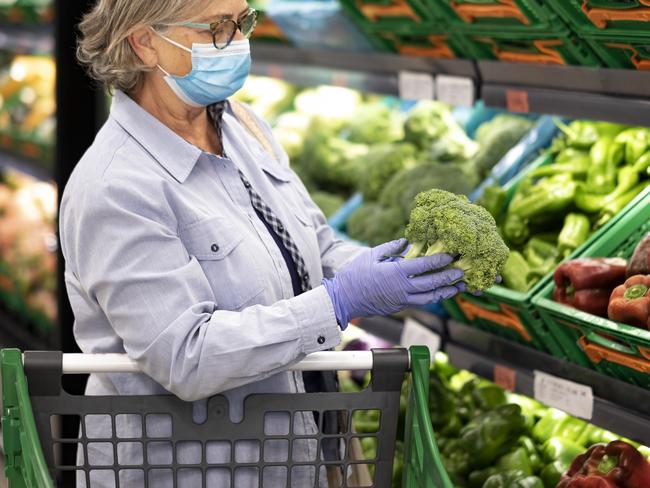 Senior woman wearing face mask and rubber gloves selects broccoli and other vegetables  in a supermarket -active elderly pensioners with trolley. retiree shopping generic