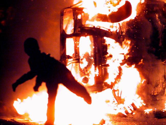 A masked youth hurls rocks at a burning vehicle on the Garvaghy road in Portadown, Northern Ireland  Mar 18, 1999, after more violence erupted following the funeral of  human rights lawyer Rosemary Nelson who was killed by a car bomb earlier this week. (AP Photo/Paul/Faith/PA)) Riots o/seas conflict fires flames illuminations silhouette vandalism generic