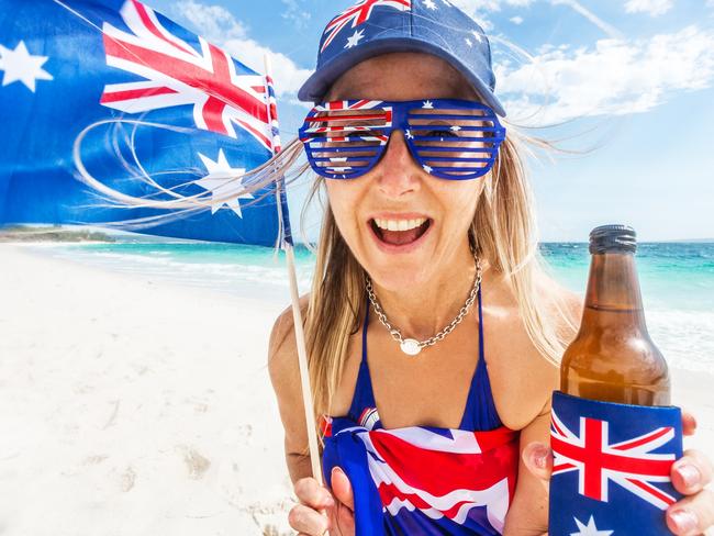 Australia Day national public holiday January 26.  Woman on beach wearing patrotic items of clothing bearing the Australian flag and she is also waving a small Australian flag and holding a drink in a cooler