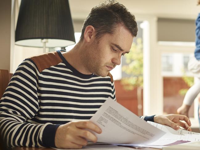 A young husband and father sits at a dining table looking through the household bills and looks worried . His wife and young daughter stand in the background looking on sympathetically .