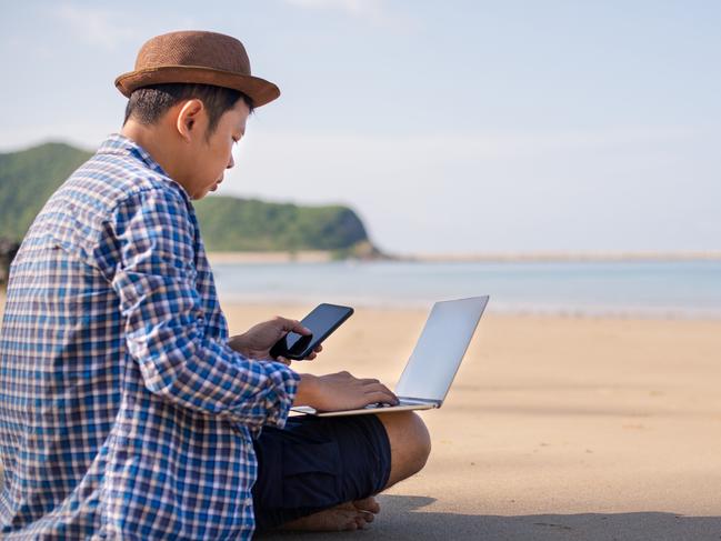 QLD JOBS - freelancer young man in a shirt and shorts and sneakers working on laptop with smartphone while sitting on the summer beach, sending mail,relax and vacation concept. Picture: Istock