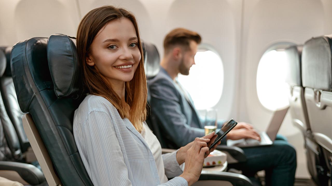 Cheerful female traveller holding smartphone and smiling while sitting in passenger chair in plane