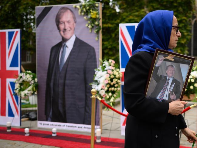 A woman carries a photograph of murdered MP David Amess walks past a row of images outside the Houses of Parliament in London. Picture: Getty Images