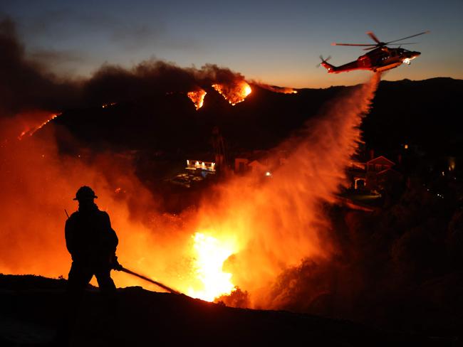 TOPSHOT - Fire personnel respond to homes destroyed while a helicopter drops water as the Palisades Fire grows in Pacific Palisades, California on January 7, 2025. A fast-moving wildfire in a Los Angeles suburb burned buildings and sparked panic, with thousands ordered to evacuate January 7, 2025 as "life threatening" winds whipped the region. Frightened residents abandoned their cars on one of the only roads in and out of the upscale Pacific Palisades area, fleeing on foot from the 770-acre (310-hectare) blaze engulfing an area crammed with multi-million dollar homes in the Santa Monica Mountains. (Photo by David Swanson / AFP)