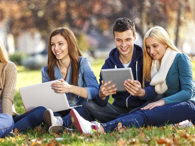 A group of cheerful university students are having a discussion in a university campus. A group consists of two boys and three girls. A girl with a long red hair in a blue shirt is holding a white laptop. A boy with brown hair is holding a digital tablet. Image: iStock