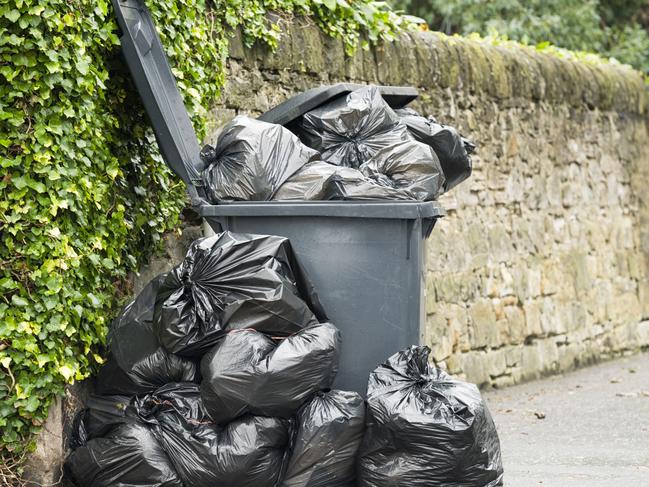 A full wheelie bin with rubbish bags overflowing onto the pavement.