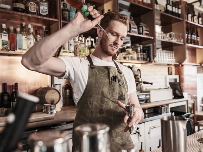 Young redhead barman working at the bar during the day. He is wearing casual clothes with bartender apron. Interior of restaurant and bar area during the day.