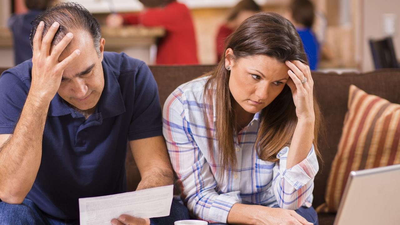 Latin descent man and woman struggle to pay their monthly bills.  They are calculating expenses versus budget income and are upset.  Many invoices, laptop on living room coffee table.  Children in kitchen background.  Mid-adult age couple.  Frustration among middle-class people.   Great imagery for election season:  home finances, recession, past due bills, mortgage, debt, stress, worry, taxes.