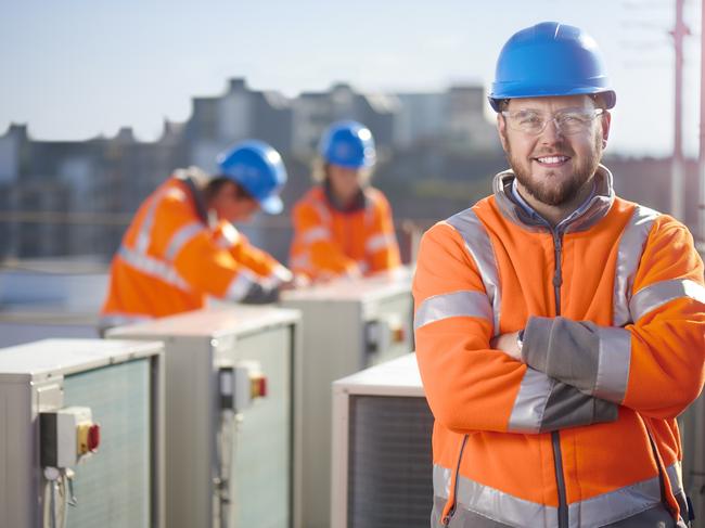 An air conditioning engineer is finishing the installation of several units on a rooftop. Two colleagues can be seen also installing units in the background. They are wearing hi vis jackets, hard hats and safety goggles. iStock image