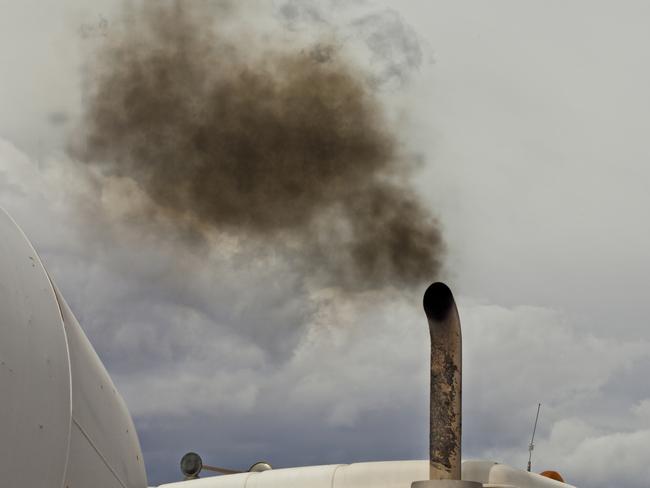 A white dump truck spews black exhaust smoke into a blue sky. Just the top of the truck is visible with the sky taking up half the image. The burning of fossil fuel by vehicles contributes to greenhouse gasses, pollution in urban city centers, and health concerns of people worldwide. Diesel fumes