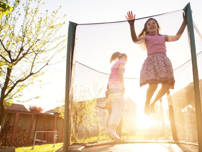 Three charming cute kids are jumping on the trampoline and the youngest girl is attempting to jump over the safety net during a beautiful sunny day.