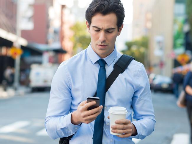 Businessman using smartphone and holding paper cup ina urban scene. Worried businessman in walking on the road and messaging with phone. Young man text messaging through cell phone while walking on the road in the city centre.