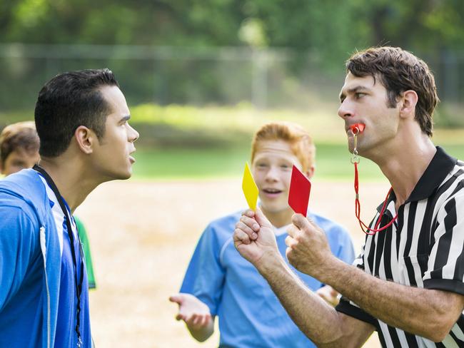 Mid adult Hispanic soccer coach argues with mid adult Caucasian referee about a penalty call. The referee has a whistle in his mouth and is showing the coach red and yellow cards. The players are in the background gesturing in unbelief. The coach is wearing a blue hoodie and the players are wearing blue jerseys. The playing field is in the background.  - Picture iStock