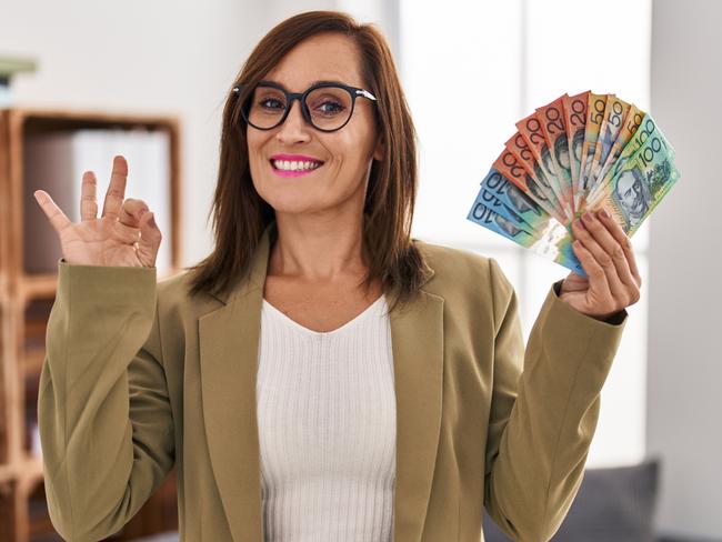 Middle age woman working at consultation office holding australian dollars doing ok sign with fingers; women female investing money generic