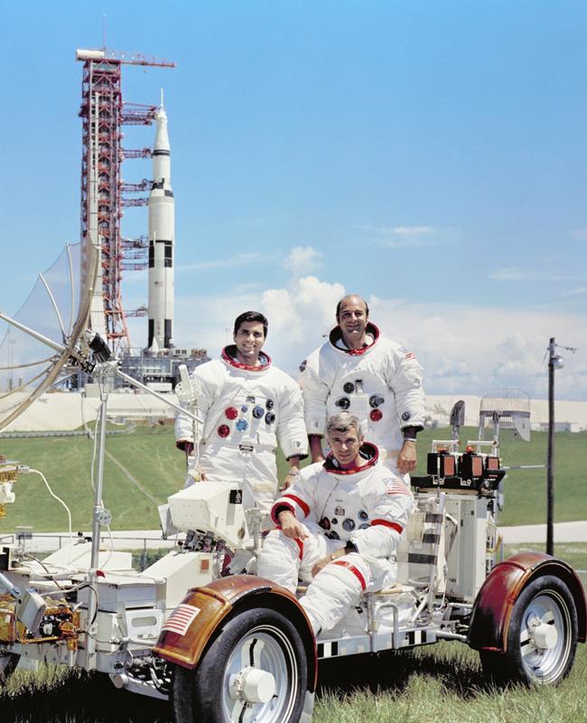 The Apollo 17 crew are photographed with a Lunar Roving Vehicle (LRV) trainer with the Saturn V Moon rocket in the background.