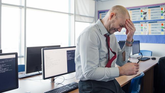 A teacher sits on the edge of a desk in an empty classroom, he is stressed