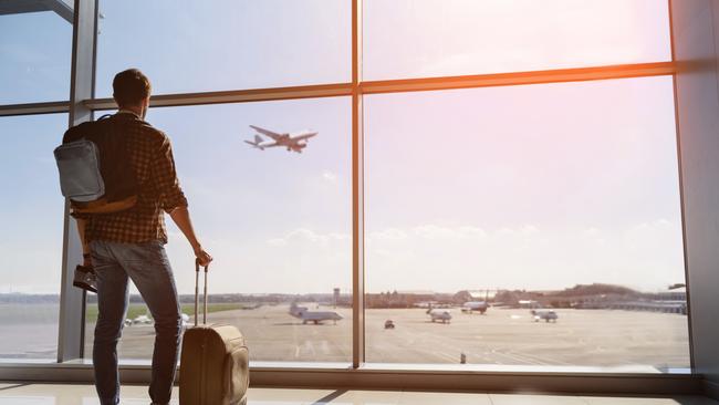 Calm male tourist is standing in airport and looking at aircraft flight through window. He is holding tickets and suitcase. Picture: iStockPaul Ewart, Tuesday Escape, travel habits