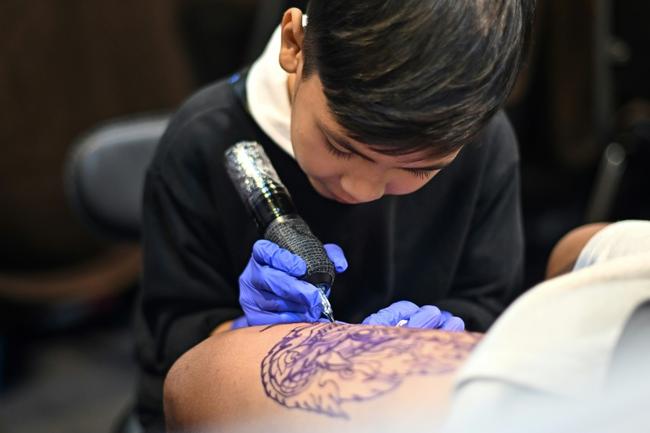 Wielding a cumbersome tattoo gun, nine-year-old Napat Mitmakorn inks the pattern of a fanged serpent on a man's upper thigh