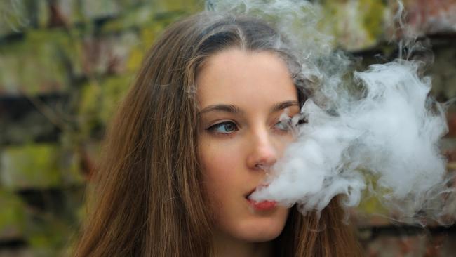 Vape teenager. Young pretty white girl in white cardigan smoking an electronic cigarette opposite destroyed brick wall on the street in the spring. Bad habit that is harmful to health.