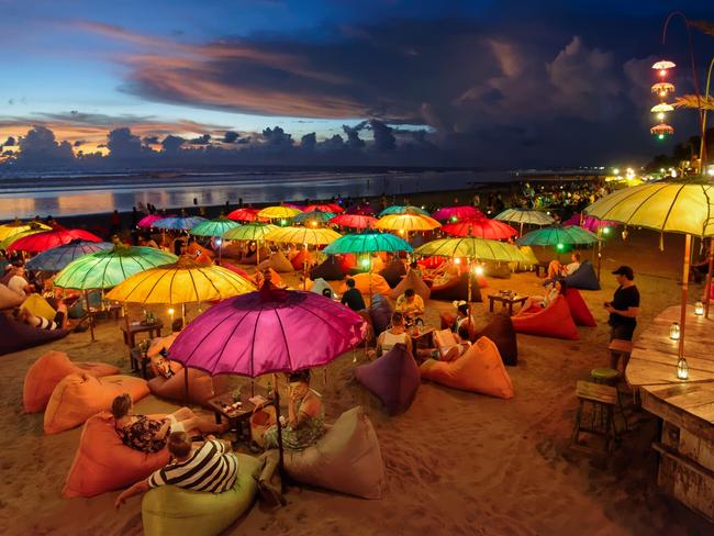 Seminyak, Indonesia.  Seminyak beach at dusk. People chilling out, having drinks, laying on puffs, under colourful lit parasolsEscape 7 January 2024Eds LetterPhoto - iStock