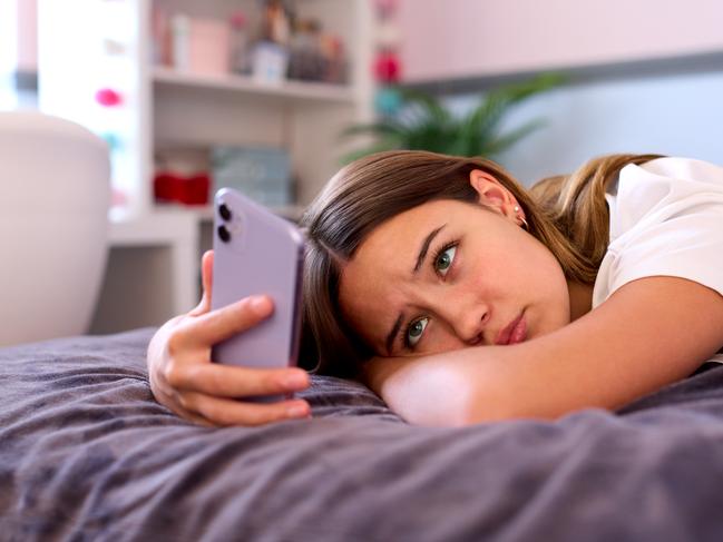 Depressed Teenage Girl Lying On Bed At Home Looking At Mobile Phone