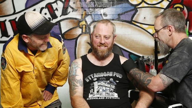 Stuart McEwan applies a bushfire commemoration tattoo to RFS volunteer Rodney McSorley, watched on by brother Darryl. Picture: John Appleyard