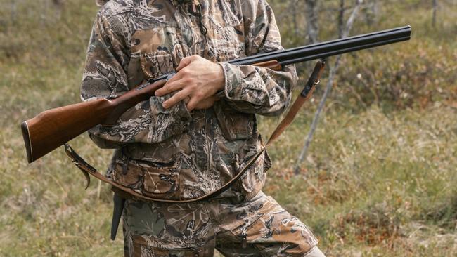 An experienced hunter in full camouflage gear steadies his rifle while navigating through a swampy area. His outfit blends seamlessly with the surrounding foliage, and the careful handling of the firearm emphasizes responsible gun control. The setting suggests a typical hunting scene, capturing a moment of anticipation and readiness in the wild.