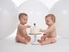 One year old twin brothers wearing diapers and eating their birthday cake. Shot in the studio with large, white balloons in the background.