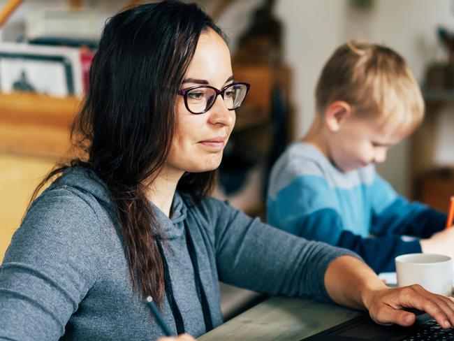 Concept of work from home and home family education. Mom and son are sitting at the desk. Business woman works on the Internet in a laptop, a child writes in a notebook.