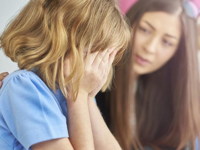 a primary school girl is sitting at her computer desk in school and crying into her hands . Her young teacher looks on  in the background , puts her arm around her and gives support .