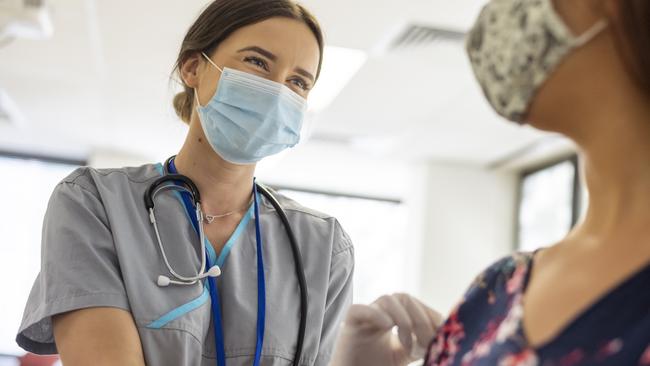 Shot focused on a kind looking nurse wearing scrubs, a stethoscope, white rubber gloves and a protective face mask. She is smiling at the nervous patient with her eyes. The nurse is prepping the patients arm before she injects her with the COVID-19 vaccine.