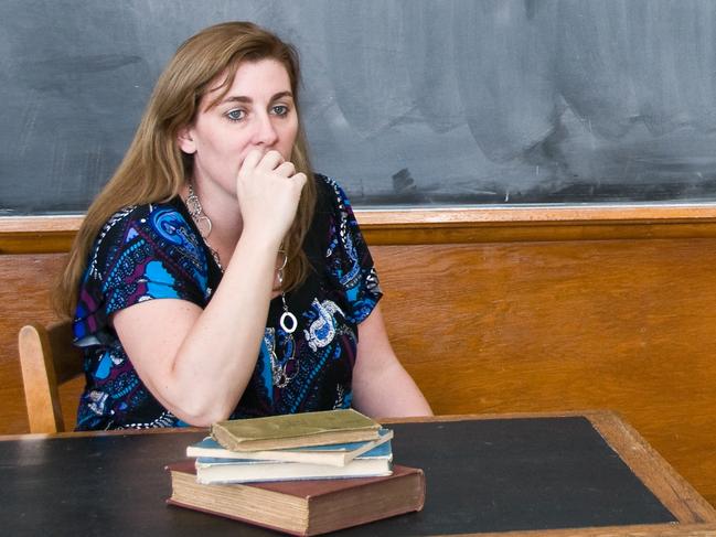 A teacher sitting at her desk looking worried with an old style blackboard behind her.