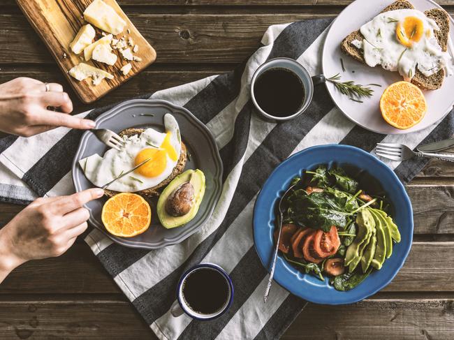 Istock image of healthy breakfast for wellbeing column in Gold Coast Eye Top view shot of breakfast table. Poached egg with bread, sliced orange and avocado are on plates. Coffee mugs, cheese plate and salad with avocado, tomatoes and arugula are on the table with a grey tea towel. Hand slicing egg on the plate.