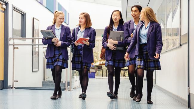 Group of teenage girls are walking down the school hall with books and laptops in their arms. They are talking and laughing as they walk and there is a female teacher using the printer.